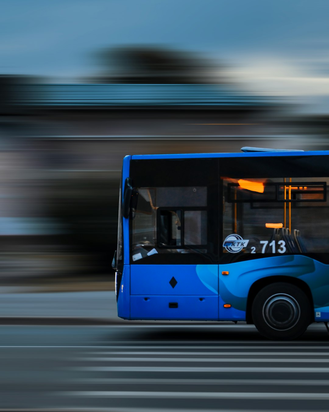 a-blue-bus-driving-down-a-street-next-to-a-tall-building-rlhi0ccnthy